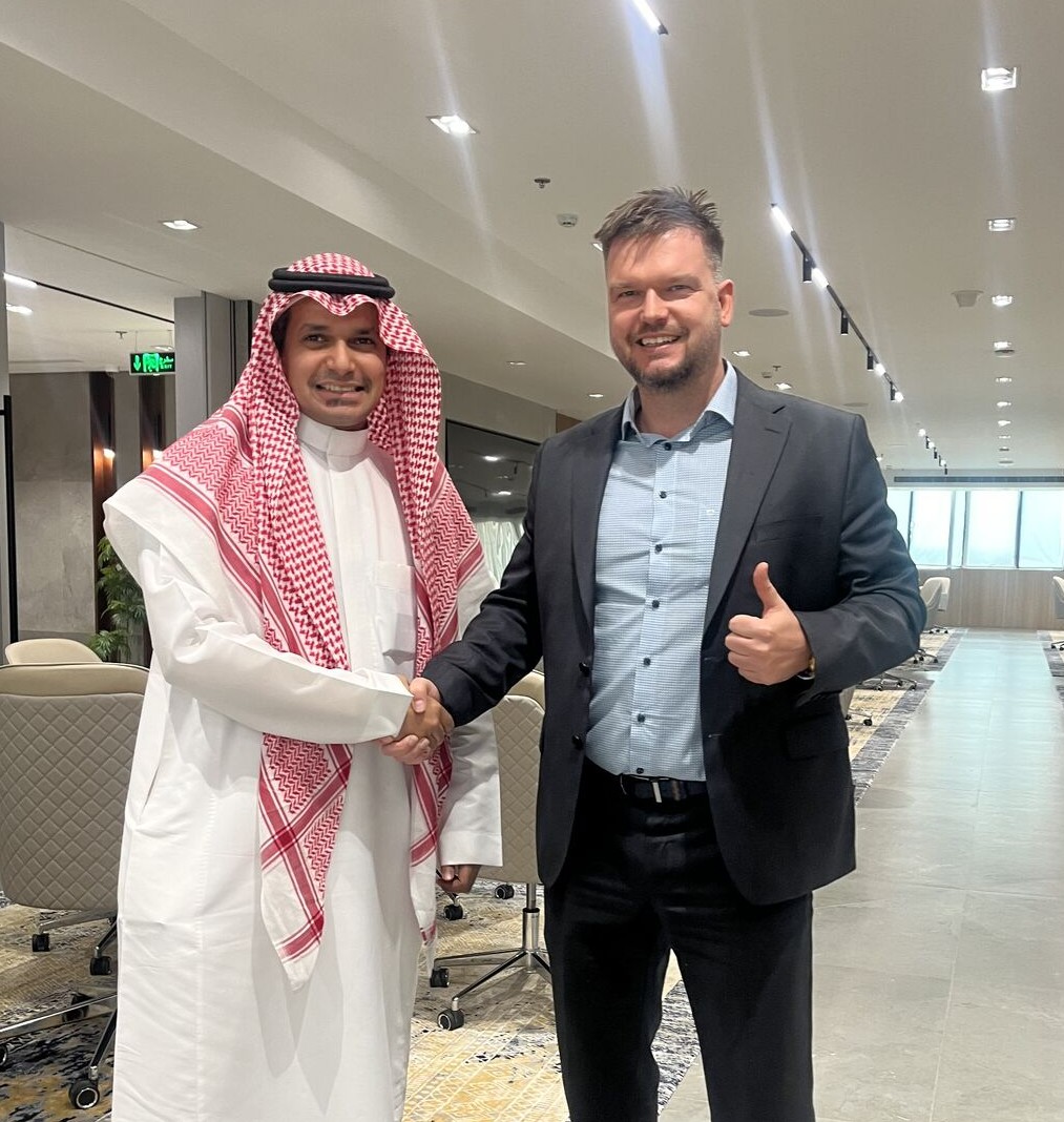Zwei Männer stehen lächelnd in einem modernen, hell erleuchteten Bürogebäude in Riad und geben sich die Hand. Der Mann auf der linken Seite trägt ein traditionelles saudisches Gewand (Thobe) und eine rot-weiß gemusterte Kopfbedeckung (Keffiyeh). Der Mann auf der rechten Seite ist der CEO von NorthIT. Er trägt einen dunklen Anzug mit einem hellblau gemusterten Hemd und zeigt mit der freien Hand einen Daumen nach oben.
